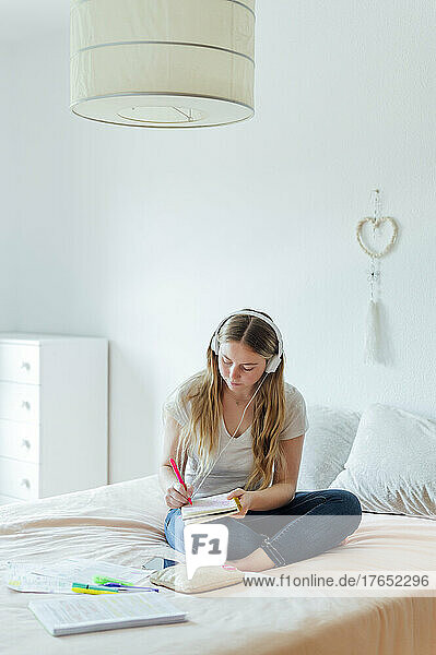 Junge Frau mit Kopfhörern macht Hausaufgaben und sitzt zu Hause im Bett