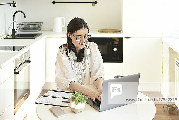 Happy freelancer using laptop sitting at table in kitchen