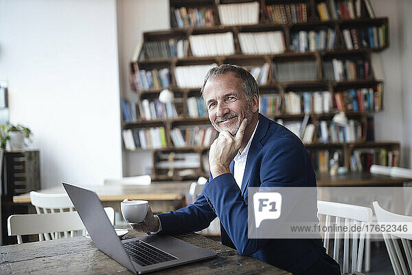 Lächelnder Geschäftsmann mit Kaffeetasse und Laptop sitzt am Tisch im Café