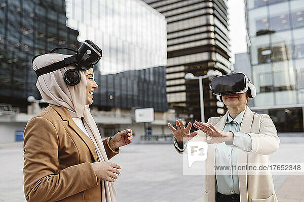 Happy young businesswoman looking at colleague wearing virtual reality simulator gesturing outside modern building