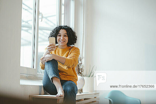 Smiling woman with mobile phone sitting on table by window at home