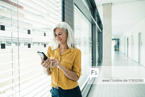Smiling businesswoman text messaging through mobile phone standing by window in office