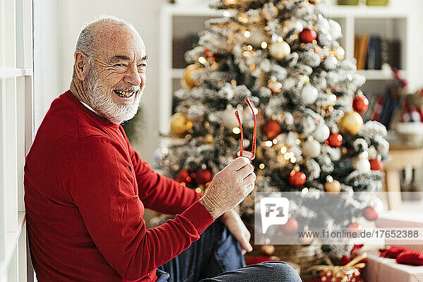 Happy senior man holding eyeglasses sitting in front of Christmas tree at home