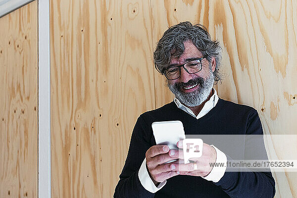 Smiling senior man text messaging through smart phone in front of wall