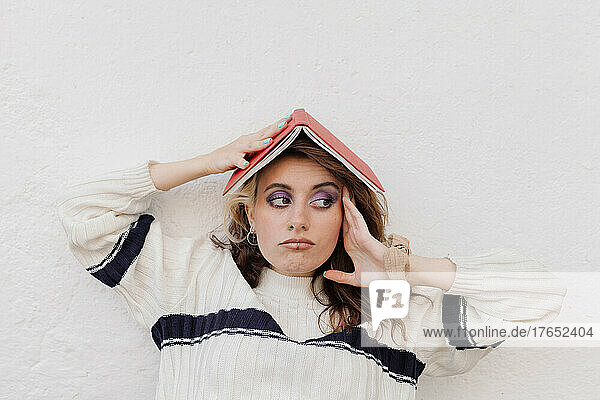 Gelangweilte junge Frau trägt Buch auf dem Kopf vor weißer Wand
