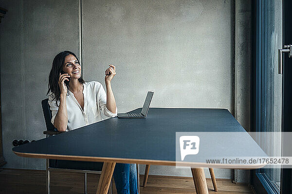 Happy woman talking on smart phone sitting at table in living room
