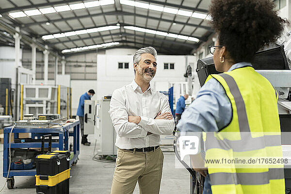 Lächelnder Geschäftsmann steht mit verschränkten Armen und diskutiert mit Ingenieur in der Fabrik