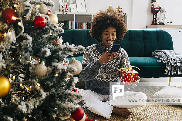 Lächelnde junge Frau fotografiert zu Hause Weihnachtsgeschenk per Handy