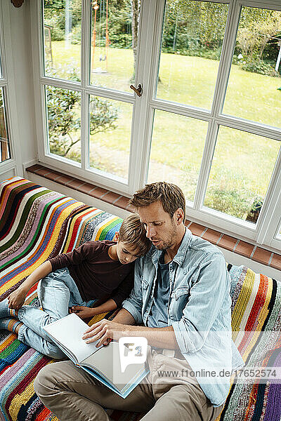 Man teaching son reading book sitting on sofa at home