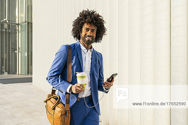 Businessman with smart phone and disposable cup walking with shoulder bag by wall