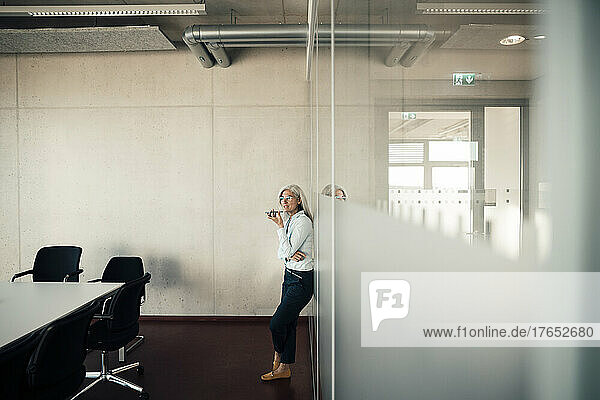 Businesswoman sending voicemail through smart phone leaning on glass wall in office