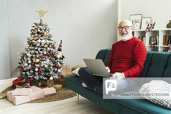 Smiling senior man with laptop sitting on sofa at home