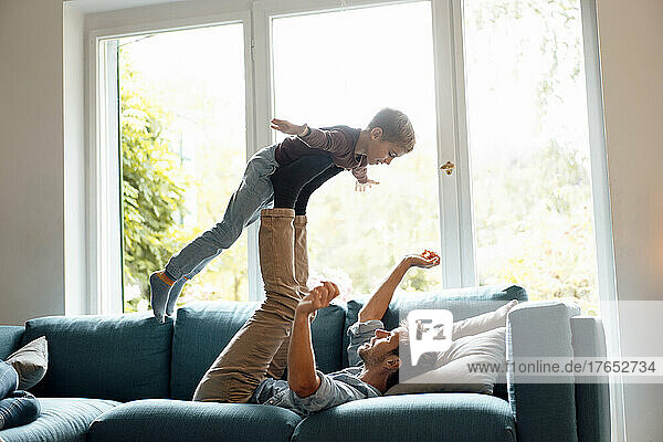 Father lifting son on legs lying on sofa in living room at home