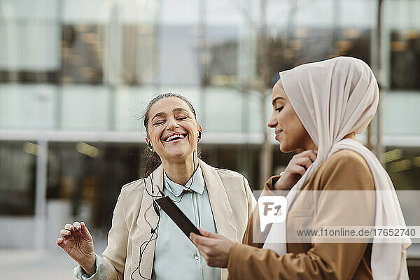 Smiling young businesswoman with tablet PC standing by happy colleague wearing in-ear headphones