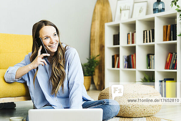 Happy freelancer with laptop talking on mobile phone at home