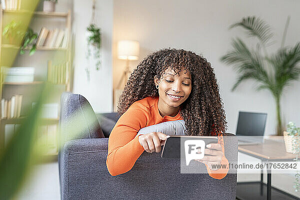 Smiling woman surfing net through mobile phone on sofa in living room
