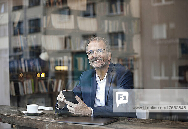 Smiling businessman with smart phone and laptop sitting by glass window at cafe