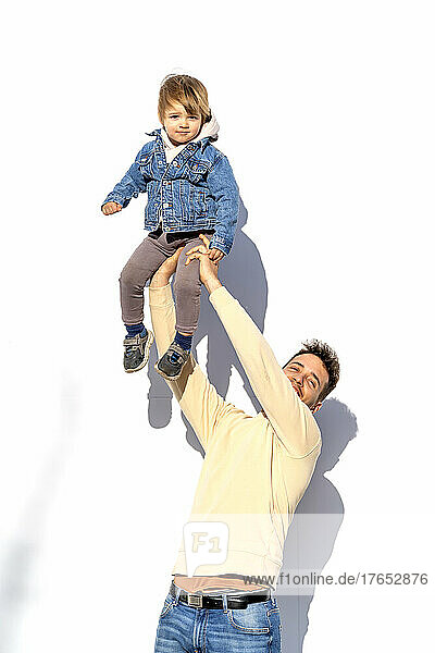 Verspielter Mann hebt seinen Sohn an der Hand hoch und steht vor einer weißen Wand