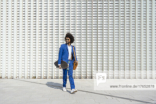Businessman holding skateboard and shoulder bag walking in front of wall on sunny day