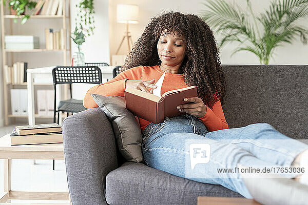 Junge Frau mit lockigem Haar liest ein Buch und sitzt zu Hause auf dem Sofa