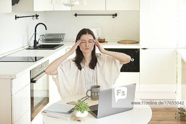 Tired businesswoman with laptop sitting at table in kitchen