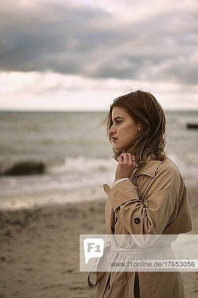 Thoughtful young woman wearing coat standing on beach at sunset