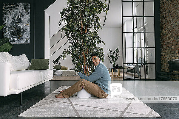 Smiling young man with eyes closed hugging houseplant sitting on carpet at home