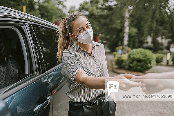 Junge Frau mit schützender Gesichtsmaske gibt einem Freund auf dem Parkplatz den Autoschlüssel