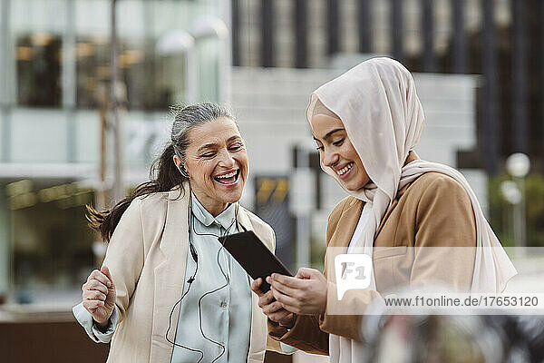 Happy businesswoman wearing in-ear headphones enjoying by colleague sharing tablet PC