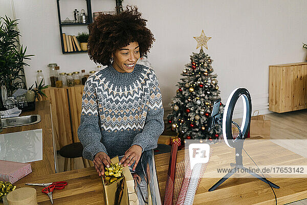 Glückliche Frau mit Afro-Frisur  die eine Geschenkbox einwickelt und zu Hause über ihr Mobiltelefon vloggt