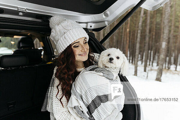 Smiling woman with cute dog wrapped in blanket sitting in car trunk