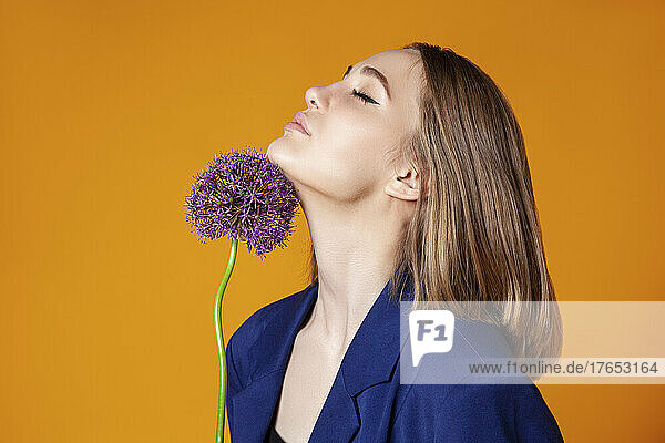 Young woman with eyes closed touching allium on chin against orange color