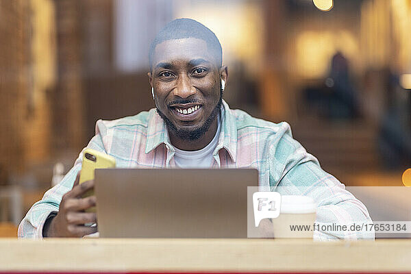 Glücklicher Mann mit Laptop und Smartphone sitzt im Café und sieht durch Glas