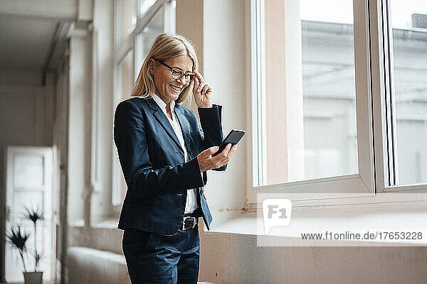 Smiling businesswoman using smart phone by window at work place