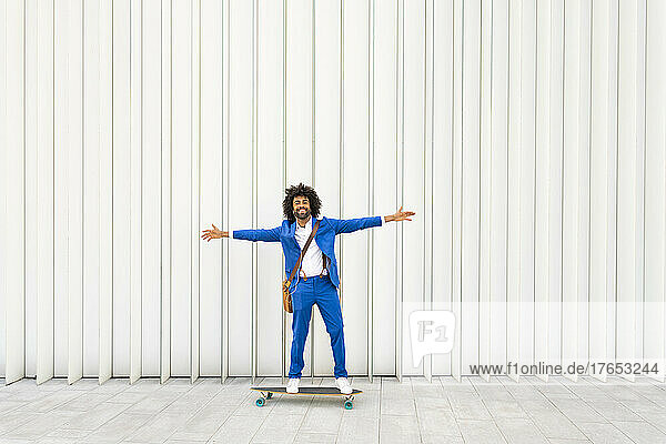 Smiling businessman standing with arms outstretched on skateboard in front of wall