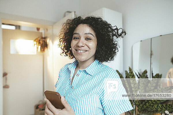 Smiling woman with smart phone standing in living room at home