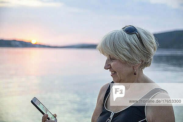 Lächelnde ältere Frau  die bei Sonnenuntergang einen Videoanruf über ihr Smartphone auf dem Seeweg tätigt