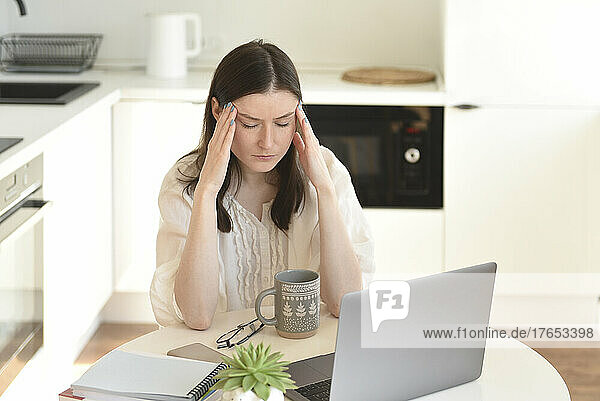 Stressed freelancer with laptop sitting at table in kitchen