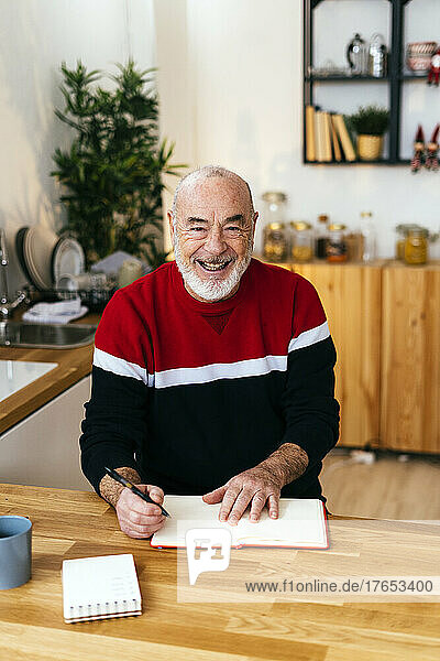 Cheerful senior man with diary sitting at table in kitchen