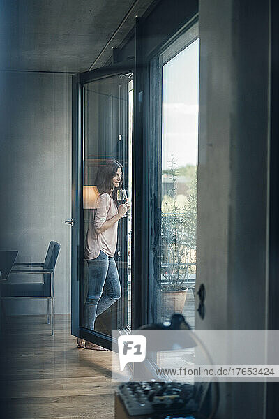 Frau mit Weinglas steht zu Hause am Fenster