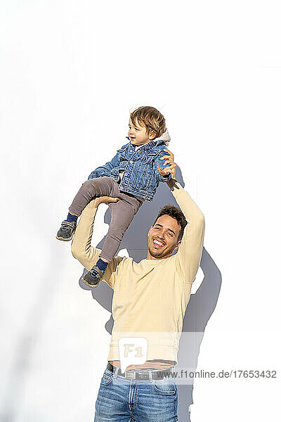 Glücklicher Mann hebt seinen Sohn an der Hand hoch und steht vor einer weißen Wand