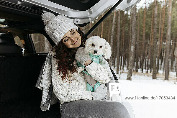 Smiling redhead woman wearing knit hat sitting with dog in car trunk