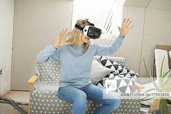 Smiling woman gesturing with virtual reality simulator sitting on sofa in attic