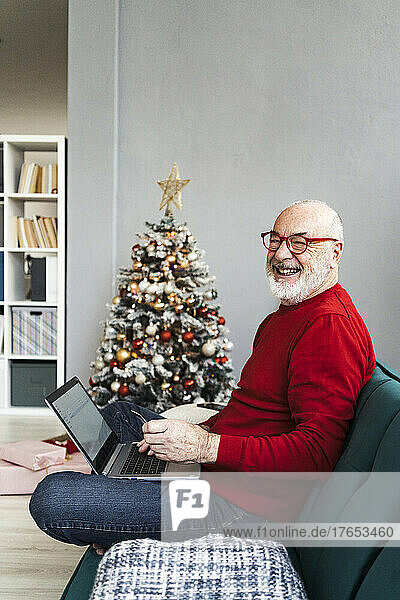 Fröhlicher älterer Mann mit Laptop sitzt zu Hause auf dem Sofa im Wohnzimmer