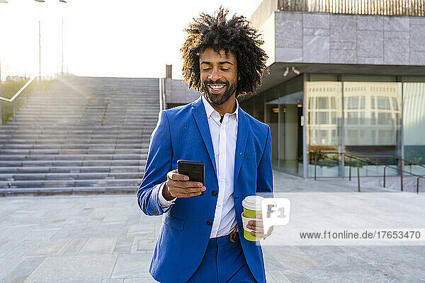 Smiling businessman holding disposable cup using mobile phone walking on footpath
