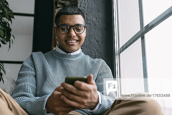 Happy man text messaging through smart phone sitting by window at home