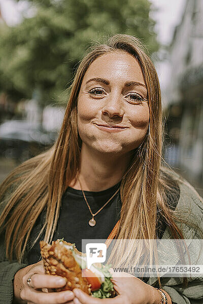Smiling young woman with puffed cheeks eating sandwich
