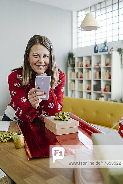 Smiling woman with mobile phone leaning on table in living room at home