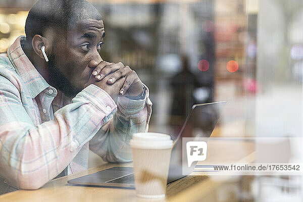 Worried freelancer with laptop sitting at cafe seen through glass