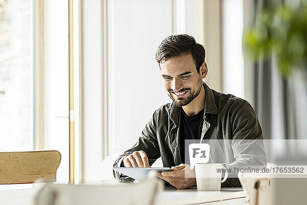 Lächelnder Mann benutzt Tablet mit Tasse auf dem Tisch zu Hause